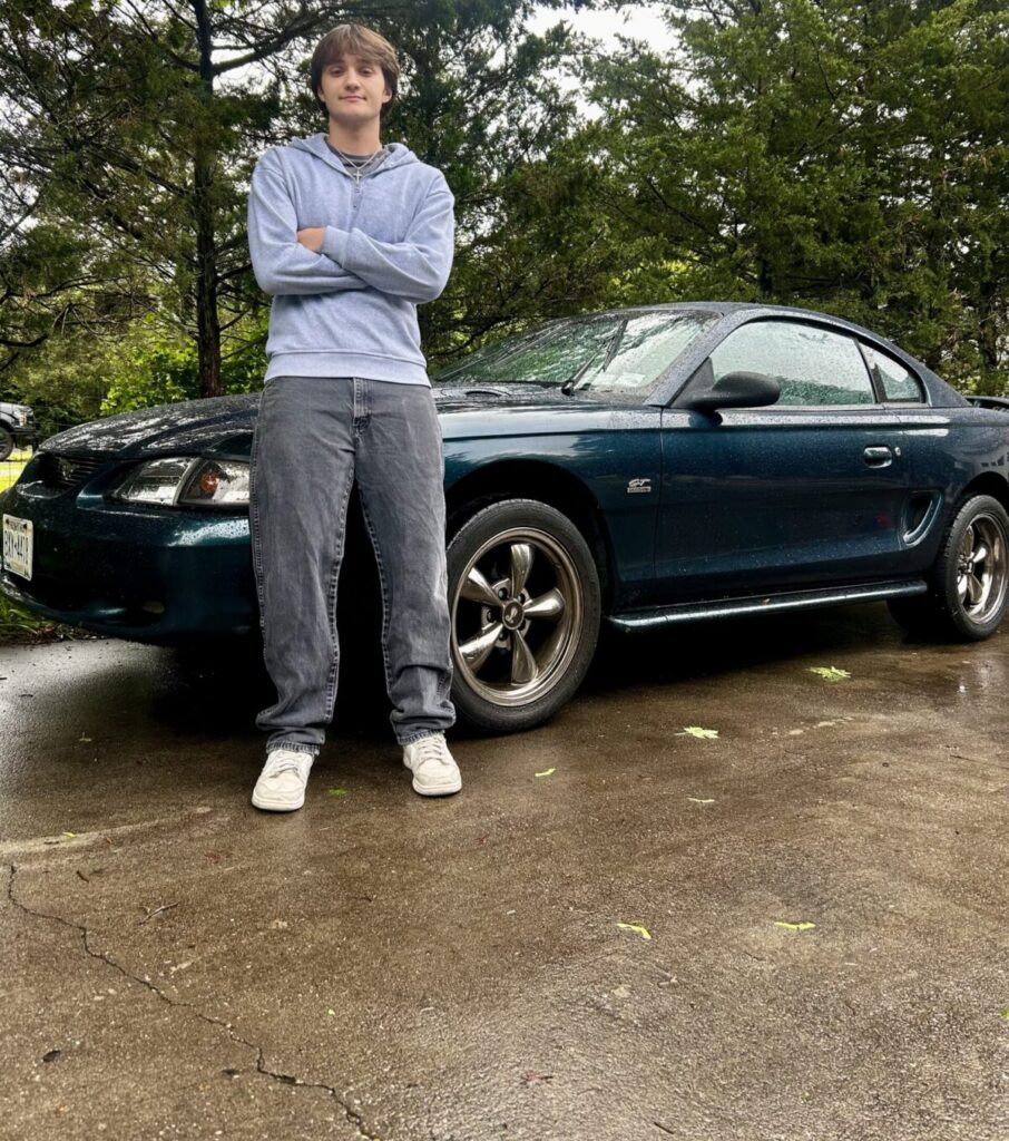 A boy stands with his arms crossed in front of a blue Mustang muscle car