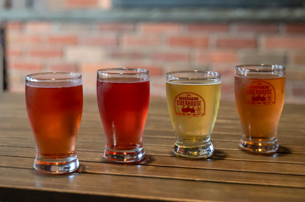 Four clear glasses, cold with condensation, are set upon a wooden slatted table top. Within them are ciders varying from blood orange to red to yellow to tangerine in color. In the background blurred slightly is exposed brick.