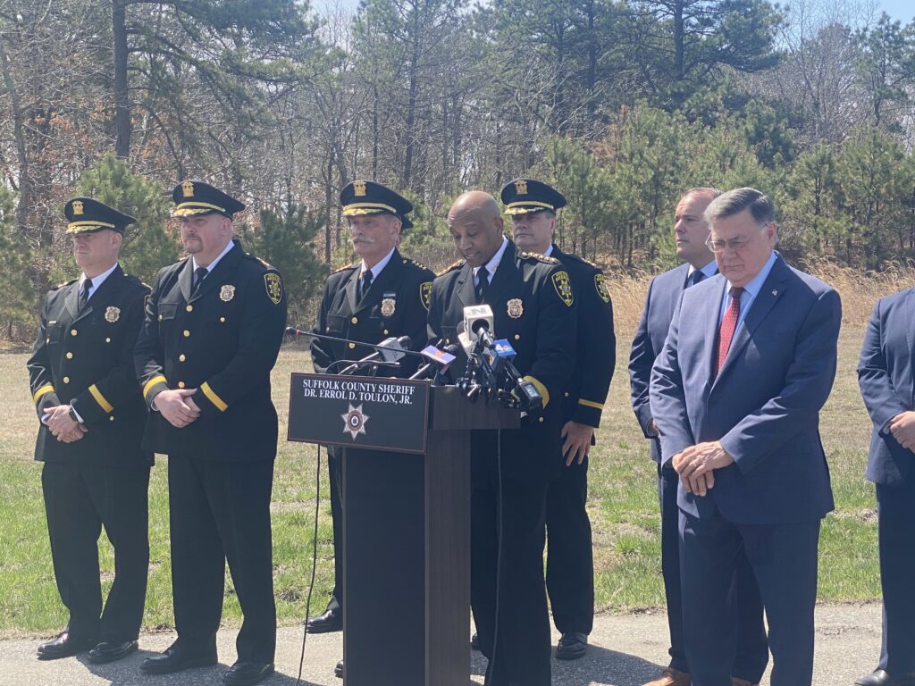 Uniformed police officers and men in suits flank the Suffolk County Sheriff, who is speaking at a podium.