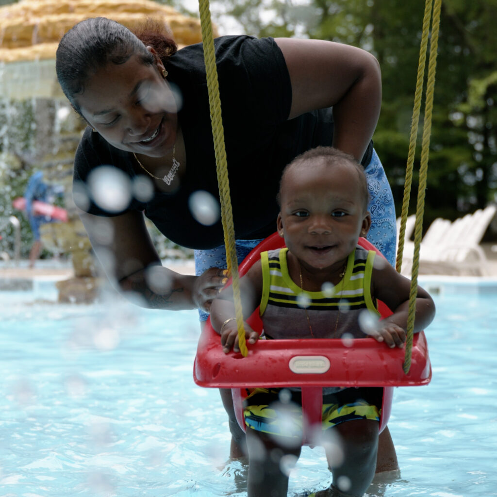 A small boy, about 2 or 3 years old sits in a red swing that sits over a shallow pool. An adult stands behind him, pushing his swing. He is smiling.