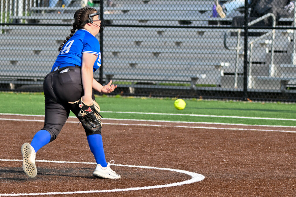 A softball pitcher in a blue jersey, black pants and blue socks pitches the ball.
